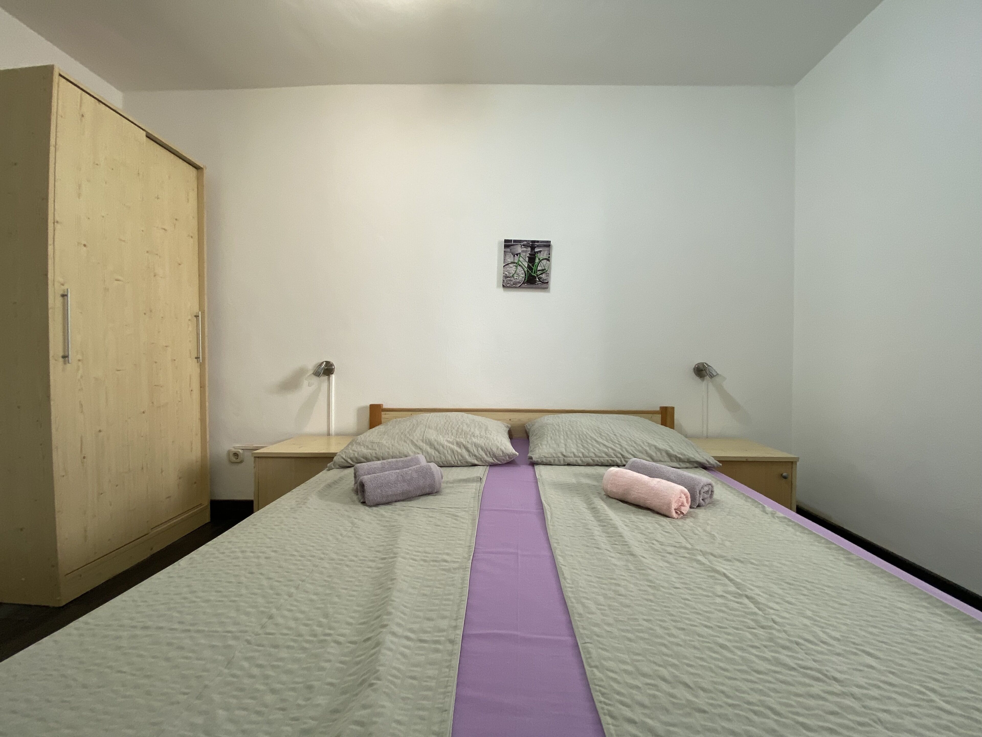 A bedroom with a bed, two nightstands, a wooden wardrobe, and a wall-mounted picture.