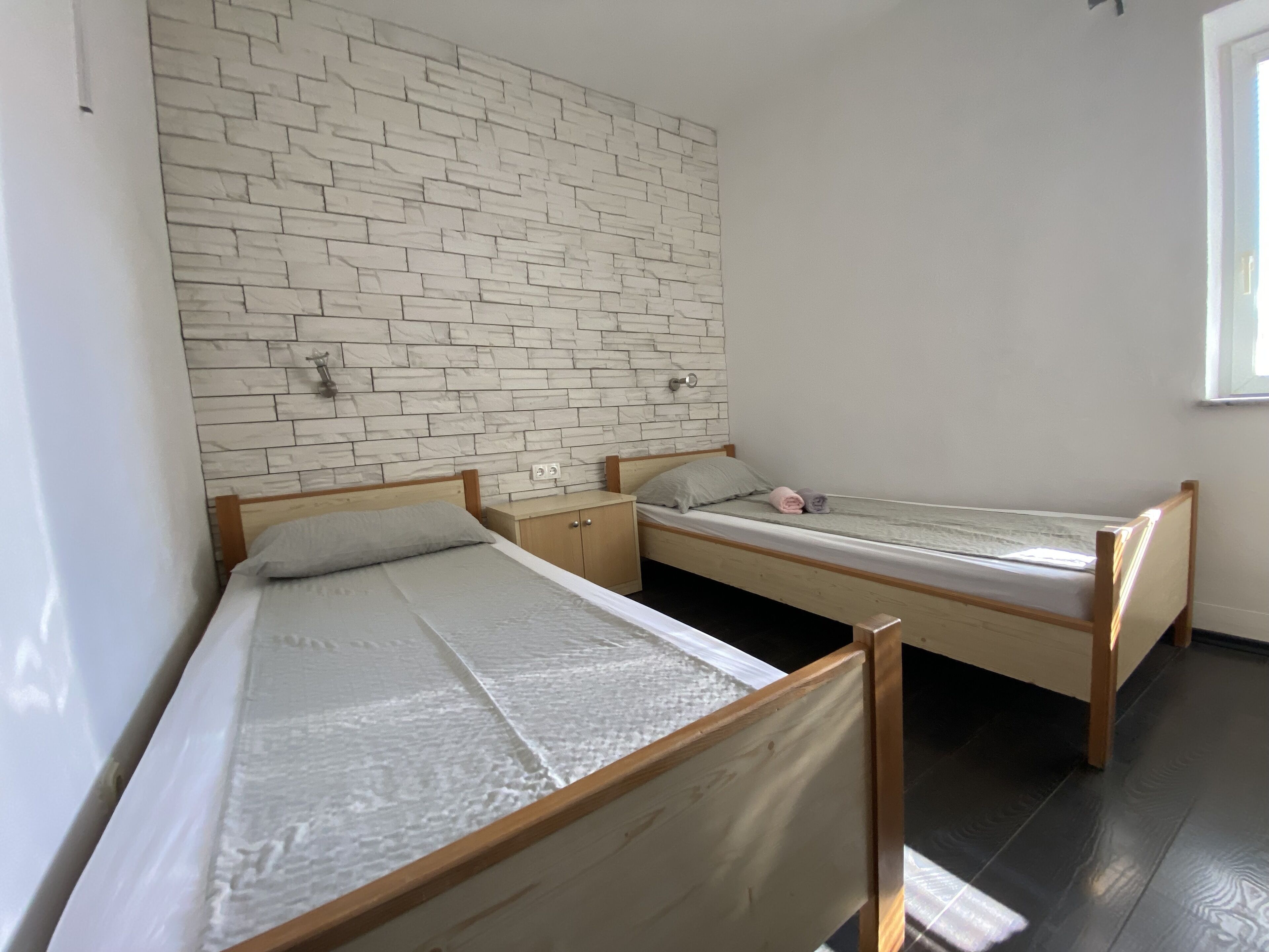 Une chambre avec deux lits en bois, un mur en briques blanches et une petite table de chevet avec une lampe.