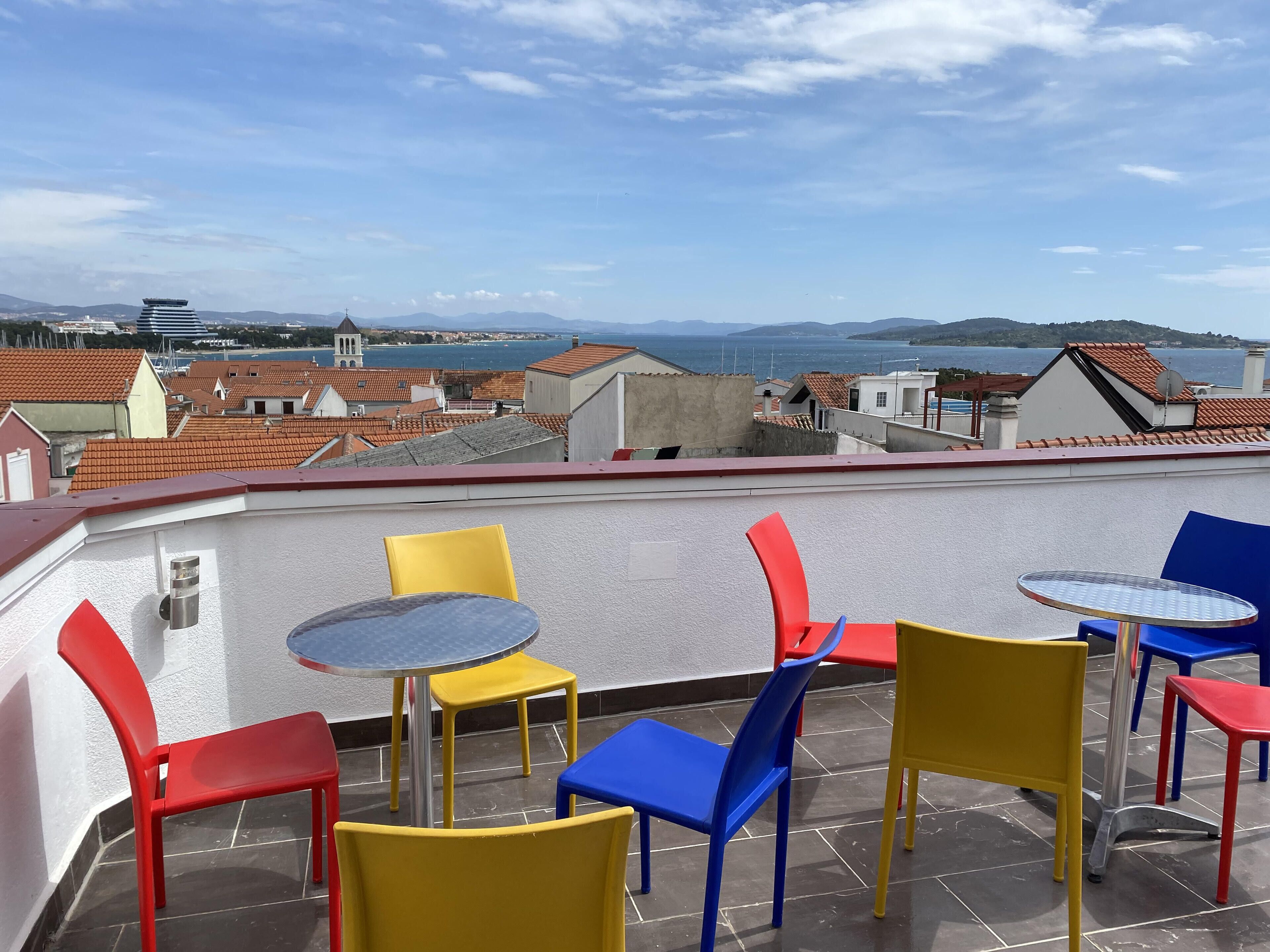Terraza en la azotea con sillas y mesas de colores, con vistas a la ciudad y al mar.