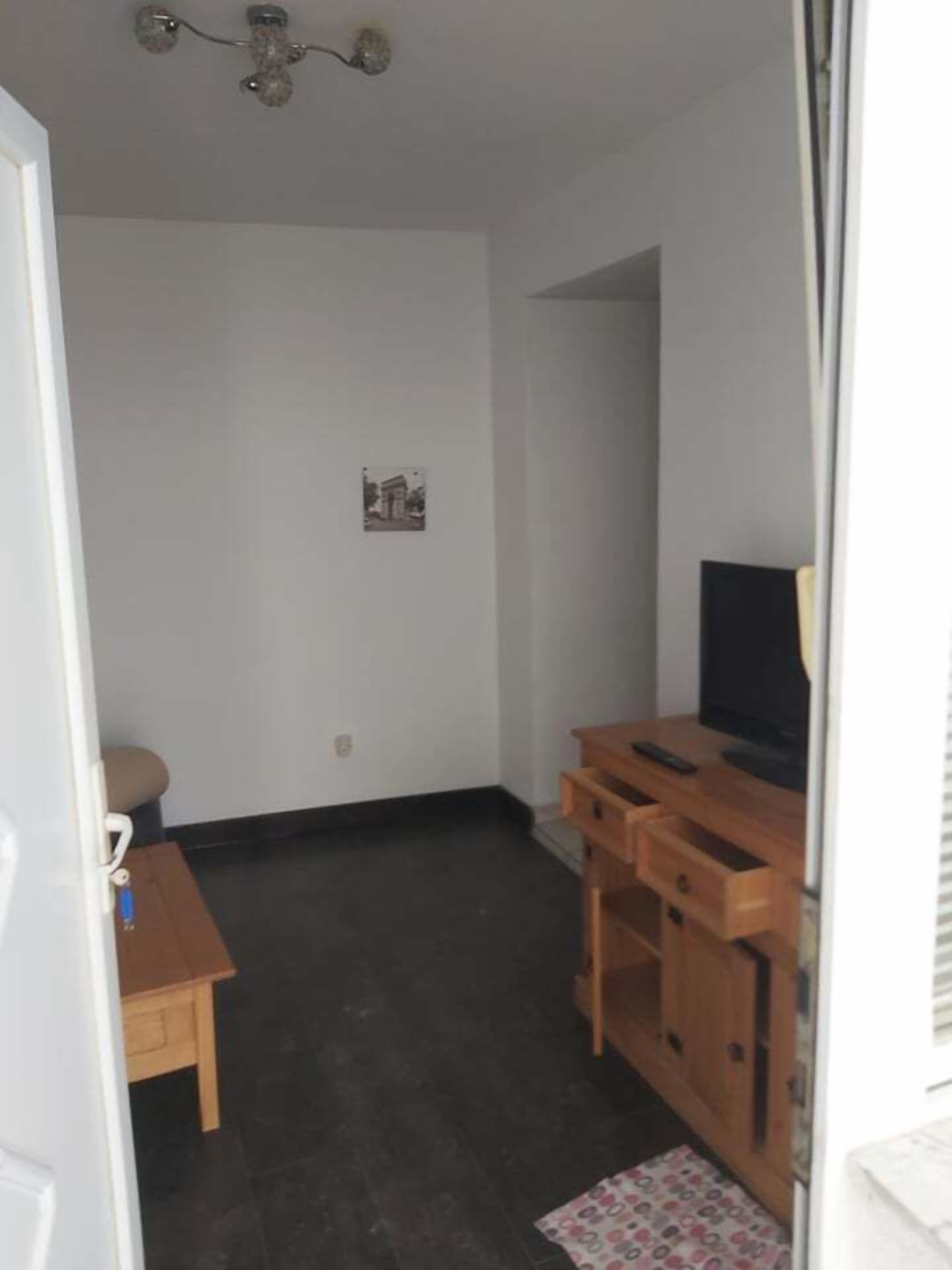 A small, sparsely furnished room with a dark floor, a wooden desk with a television, a small table, and a wall-mounted picture.