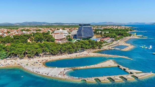 Vista aérea de una ciudad costera con un edificio moderno destacado, una playa y aguas azules cristalinas.