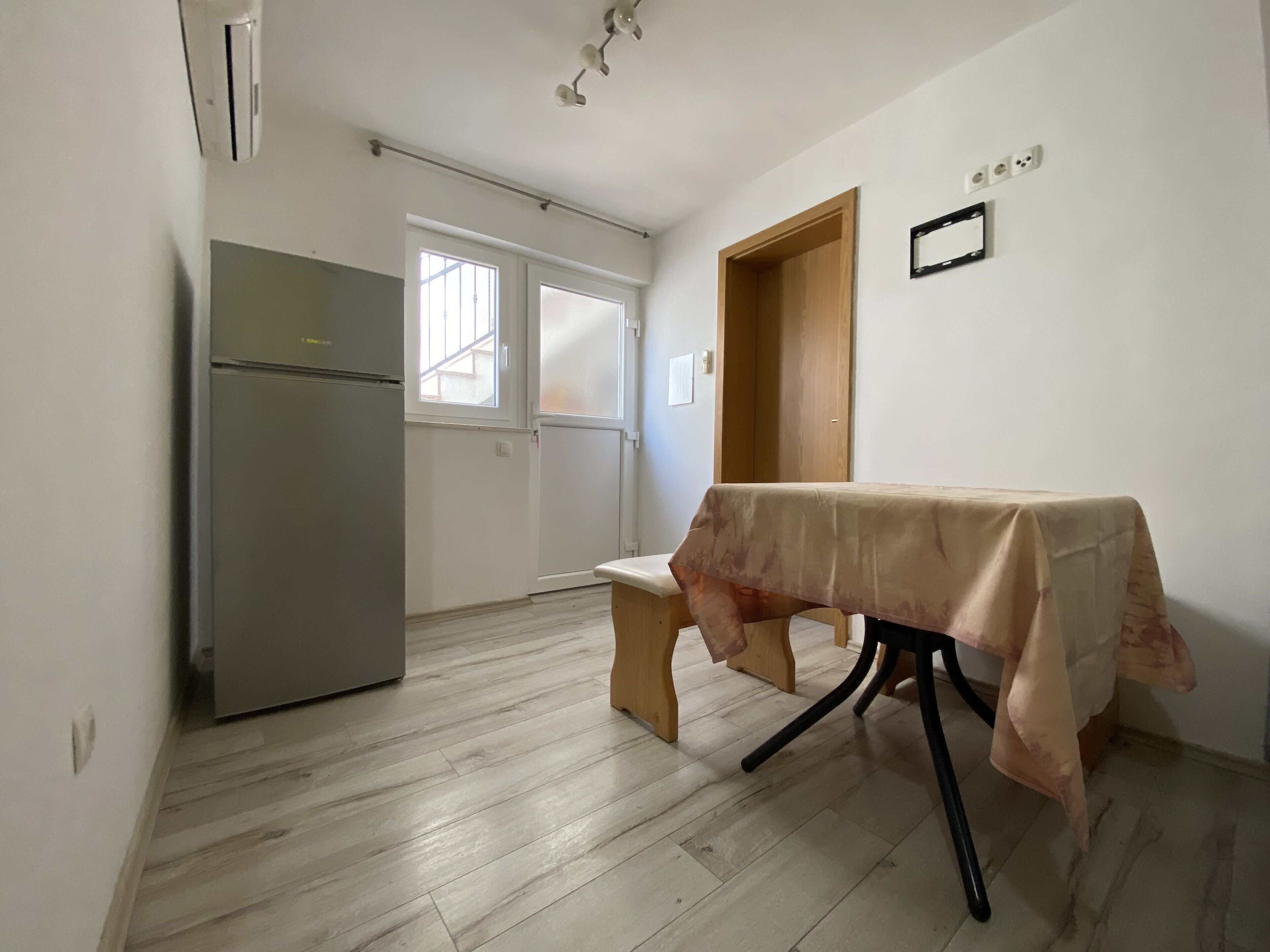 A small, open-plan kitchen and dining area with a refrigerator, a wooden bench, and a table covered with a cloth.