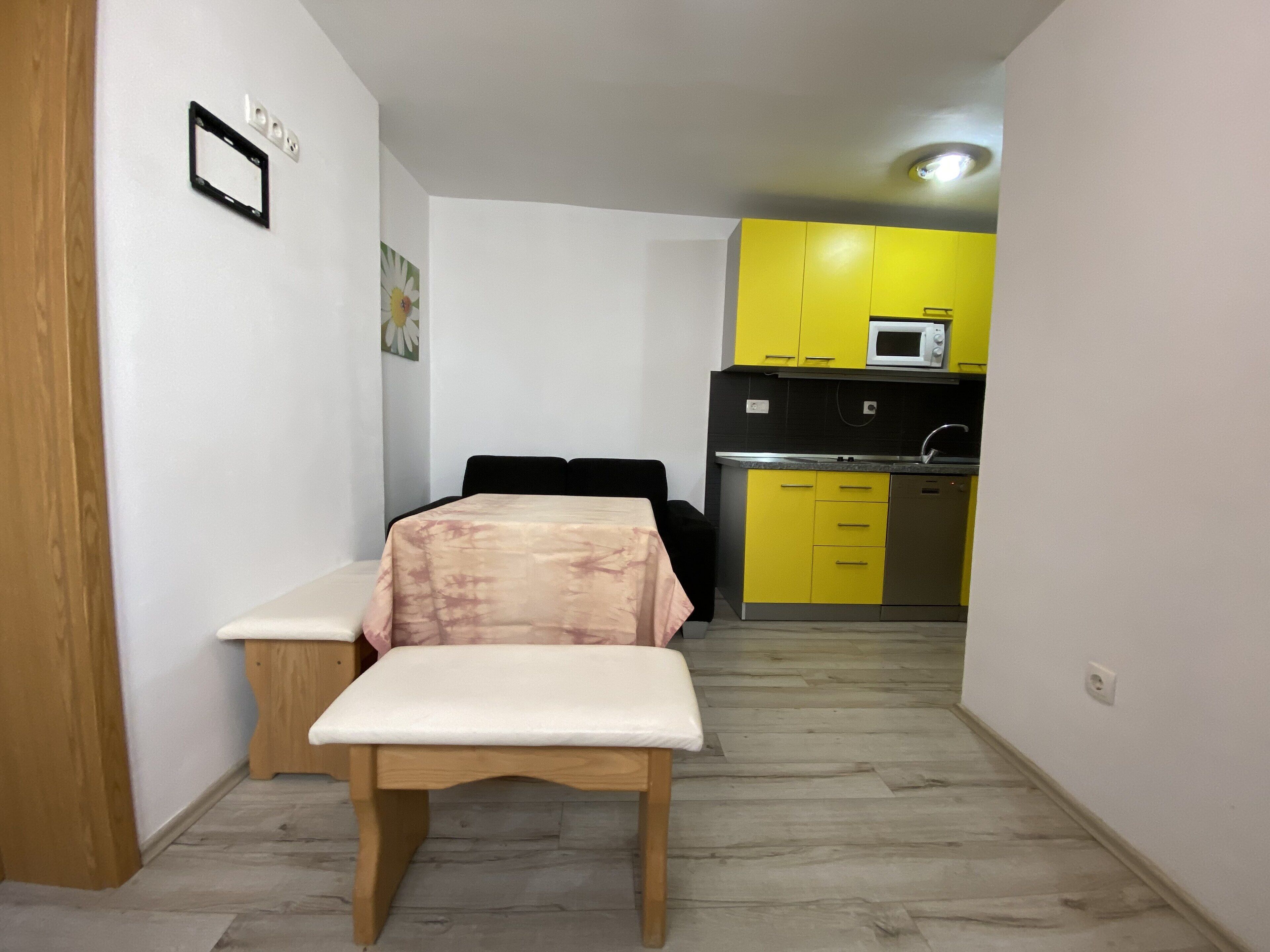 Quarto compacto com uma cama, uma mesinha e uma cozinha compacta com armários amarelos e um micro-ondas.