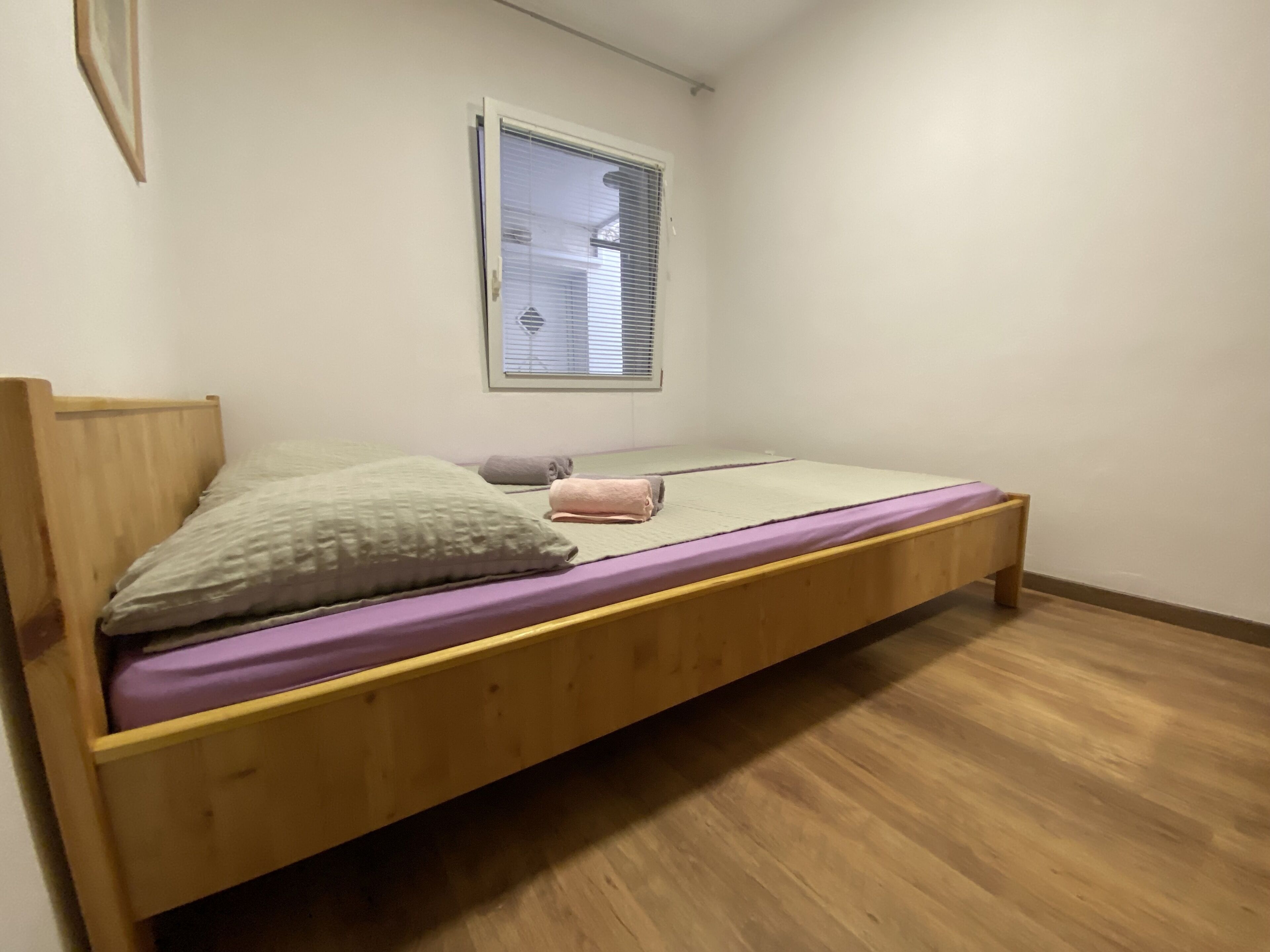 Una cama individual con estructura de madera, una ventana con persianas y una pared blanca lisa.
