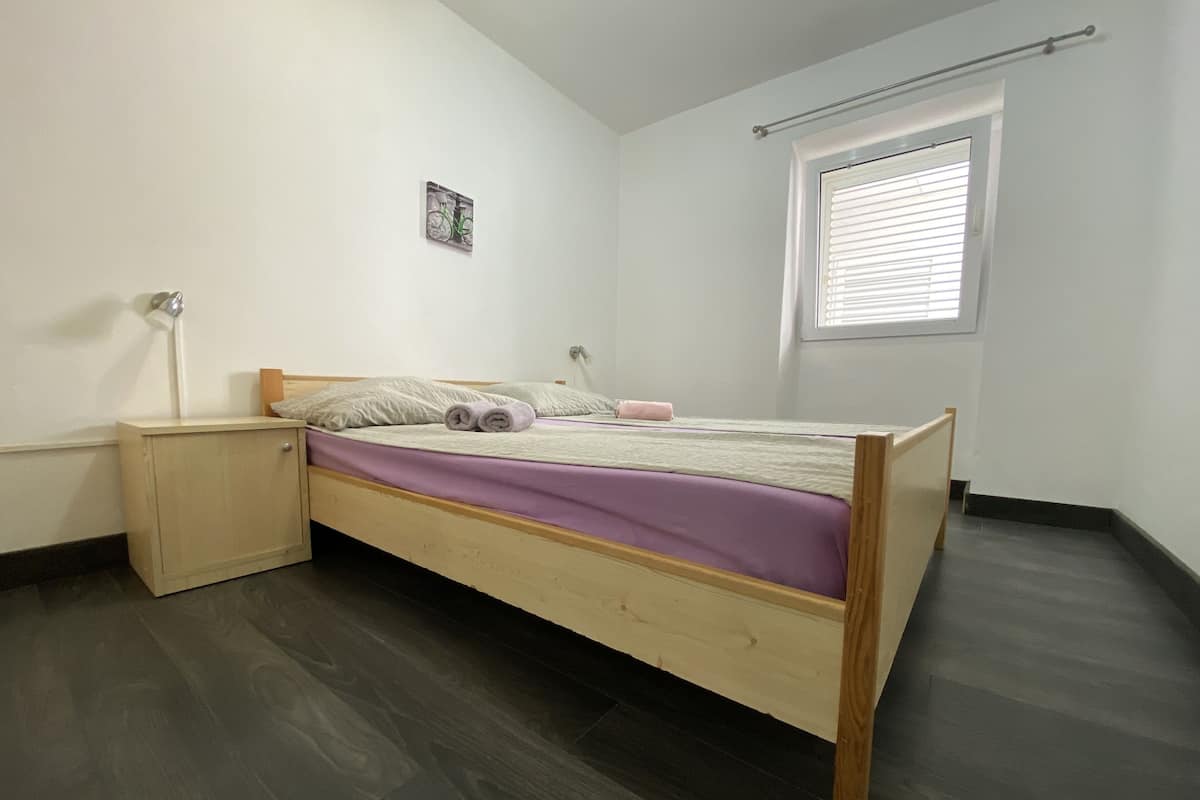 A single bed with a wooden frame, a bedside table, a wall-mounted lamp, a window with blinds, and a small picture on the wall.