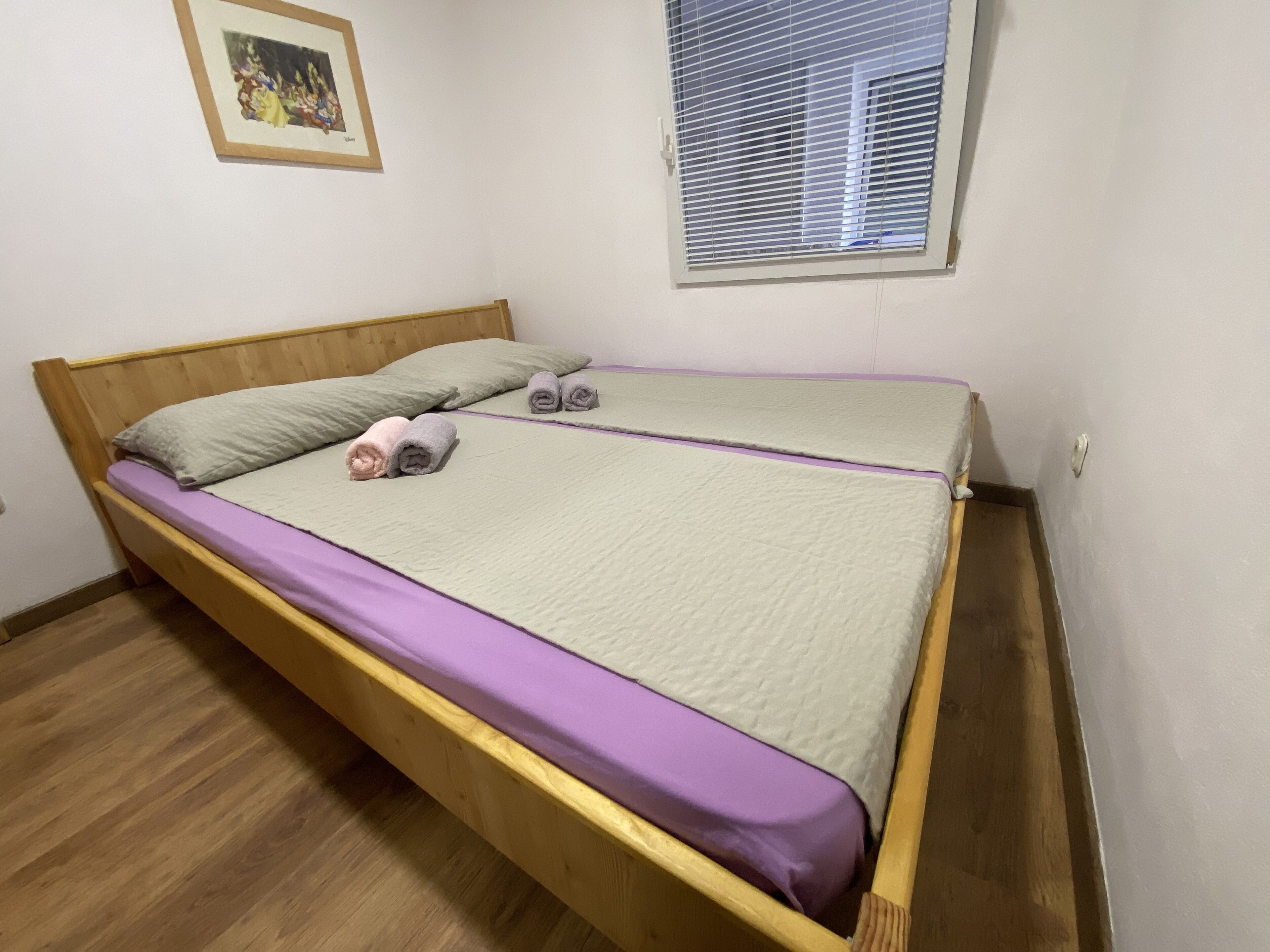 A single bed with a wooden frame, a purple mattress cover, and two rolled towels.
