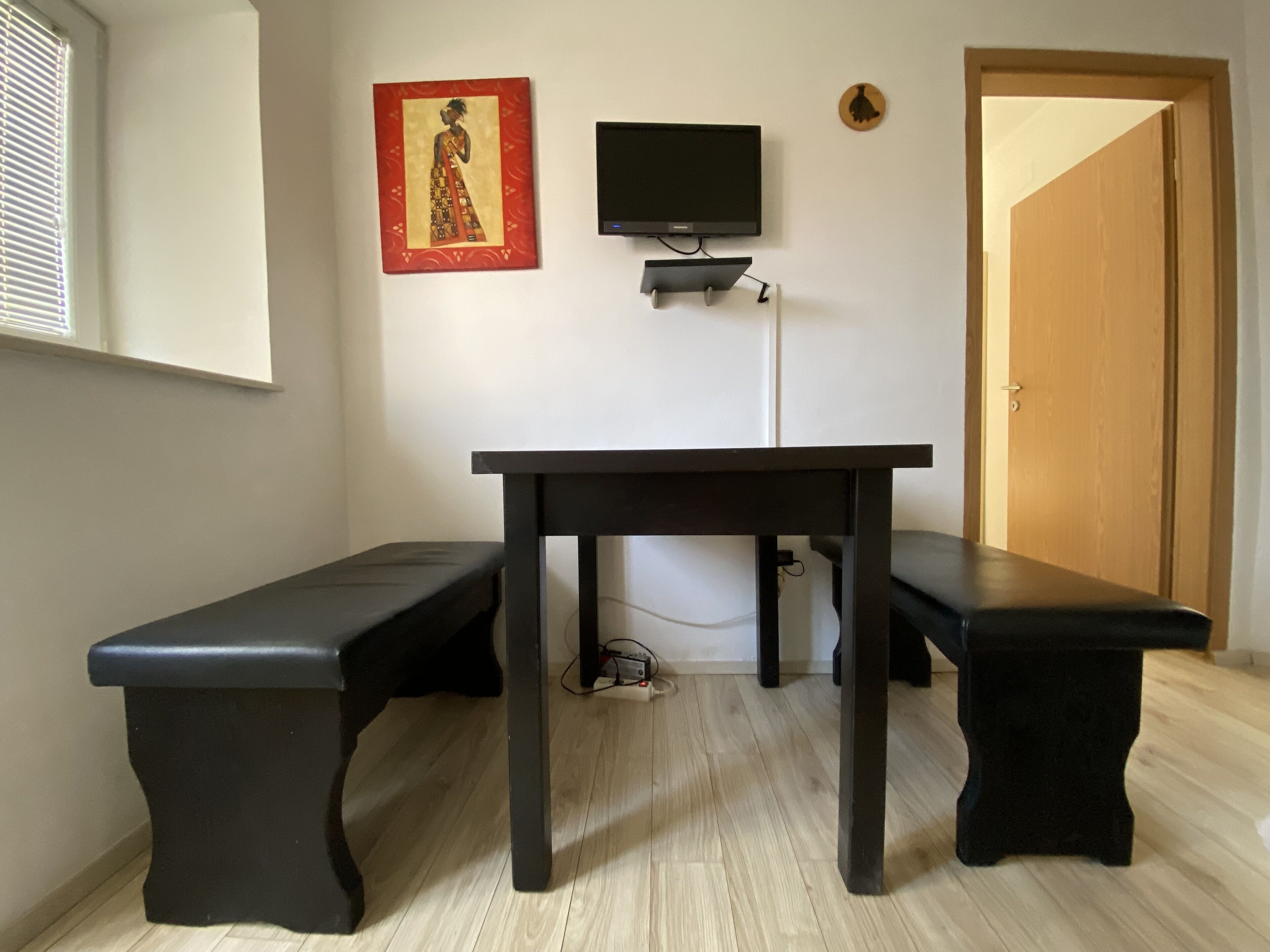 A room with a wooden floor, a black table with two benches, a television mounted on the wall, and a door.