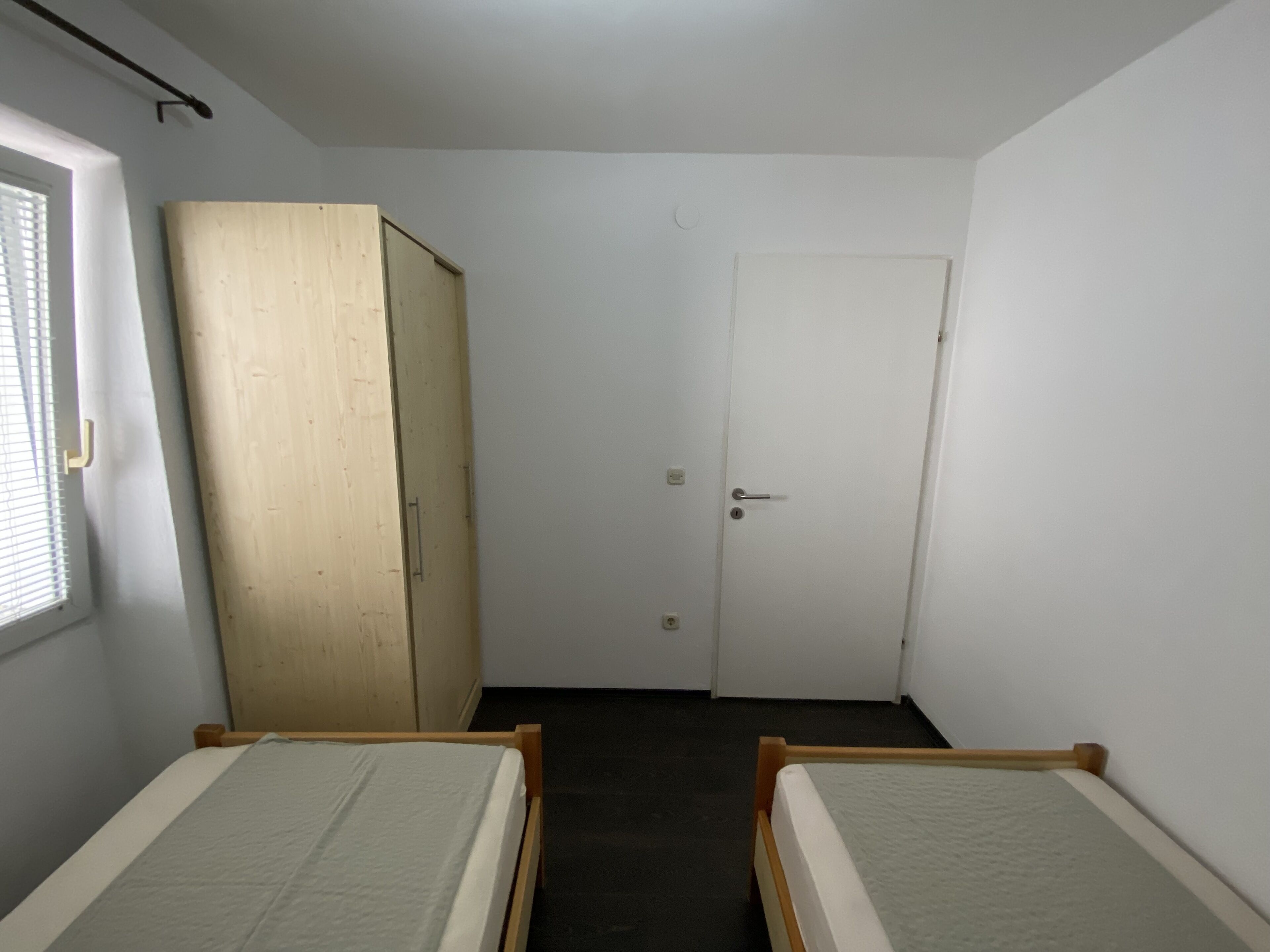 Une petite pièce étroite, avec deux lits simples, une armoire en bois et une porte blanche.