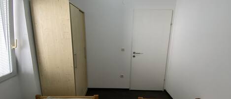 Une petite pièce étroite, avec deux lits simples, une armoire en bois et une porte blanche.