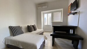 Un dormitorio con dos camas, un banco negro, un televisor montado en la pared y una ventana con persianas.