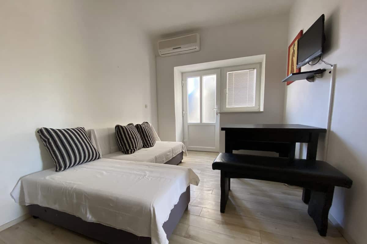 A bedroom with two beds, a black bench, a TV mounted on the wall, and a window with blinds.