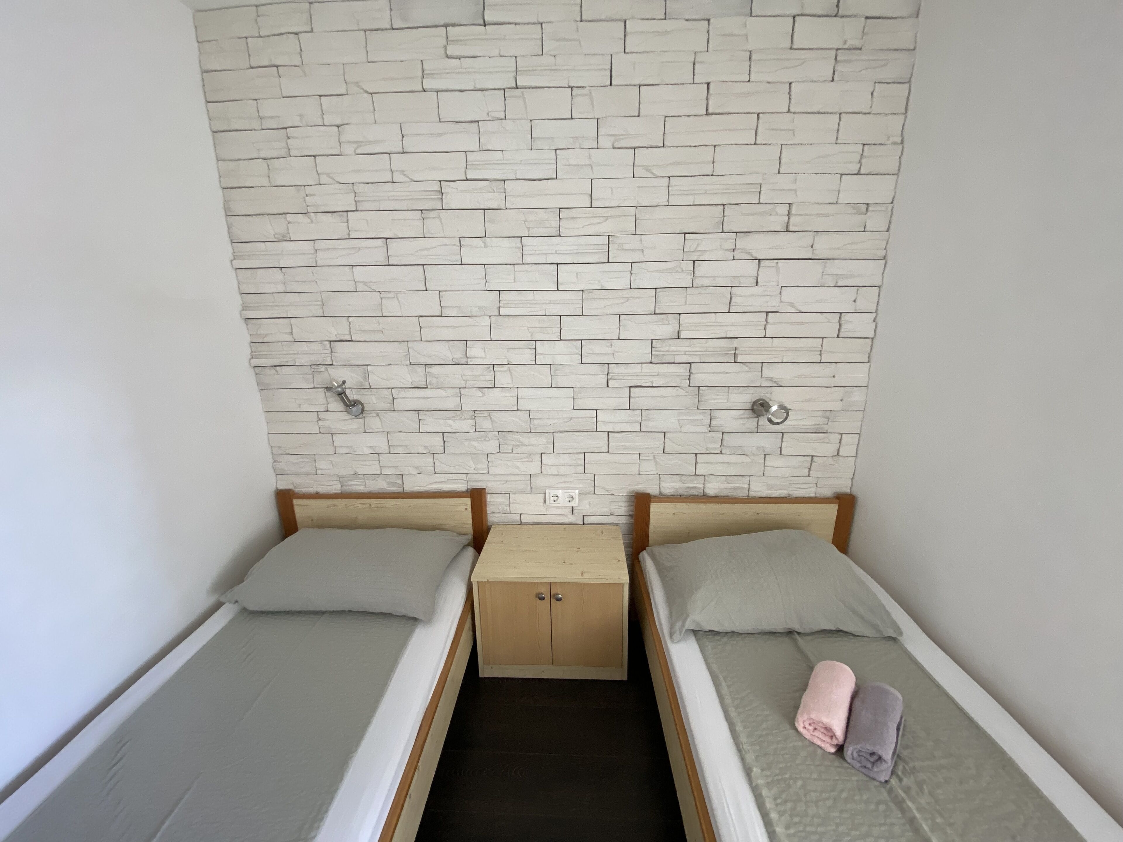 Ein Zimmer mit zwei Einzelbetten, jeweils mit hölzernen Kopfteilen und einem kleinen Nachttisch. Die Wände sind mit einer weißen Ziegeloptik versehen.