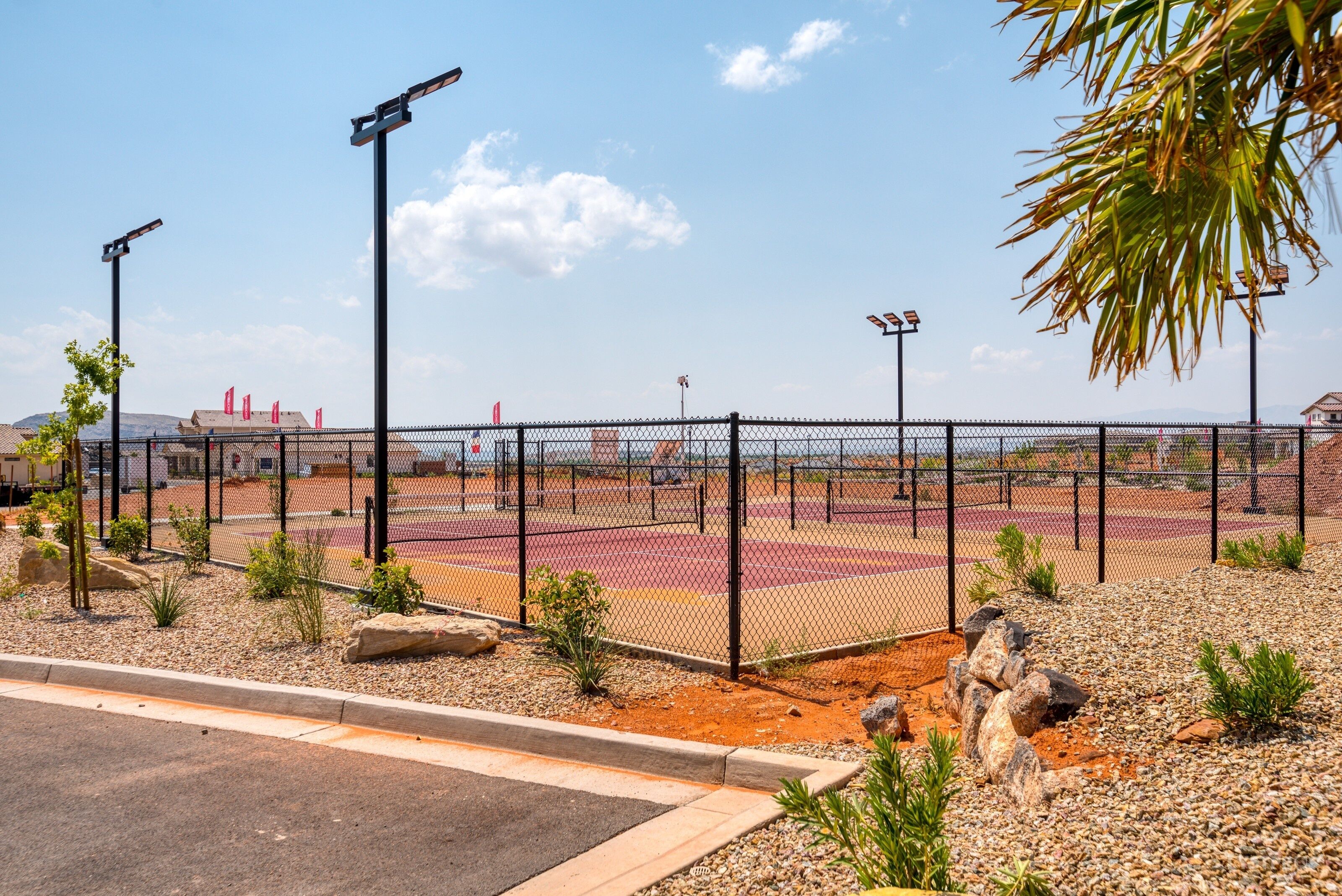 A fenced sports court with a gravel path and small plants, surrounded by a fence and street lamps.