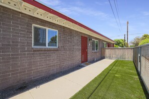 A brick building with a red door, a window, and a small concrete pathway leading to a fenced area with artificial grass.