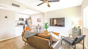A modern living room with a sofa, a dining table, and a TV. There is a ceiling fan, a kitchen in the background, and a small cabinet. - Relaxing Desert Botanical 2 Bedroom In Biltmore (Phoenix)