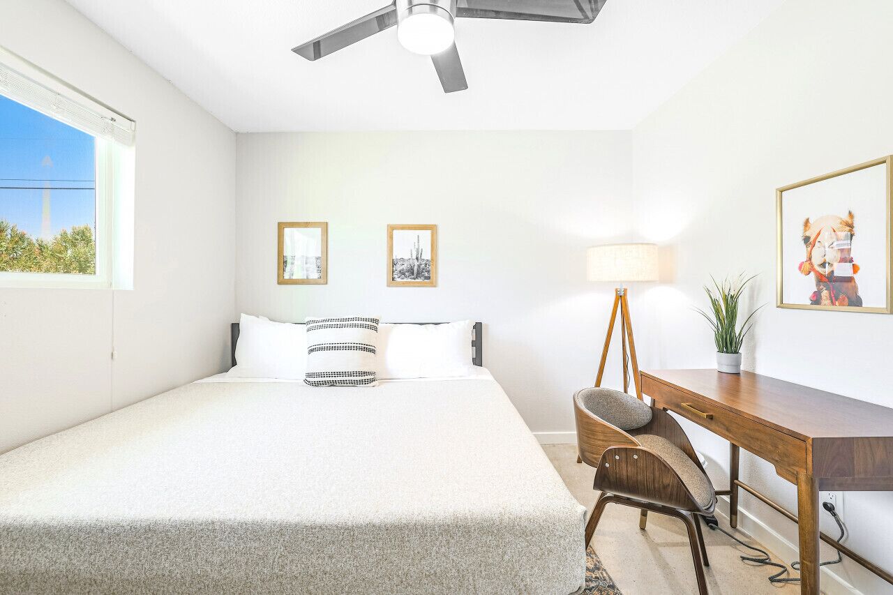 Un dormitorio con cama, escritorio, silla, lámpara y dos cuadros en la pared.