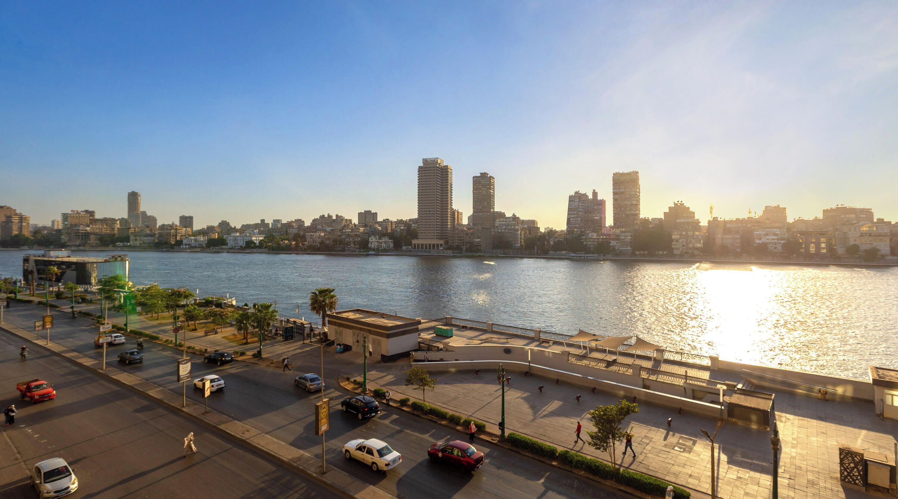A waterfront cityscape with modern buildings, a river, and a pedestrian walkway.