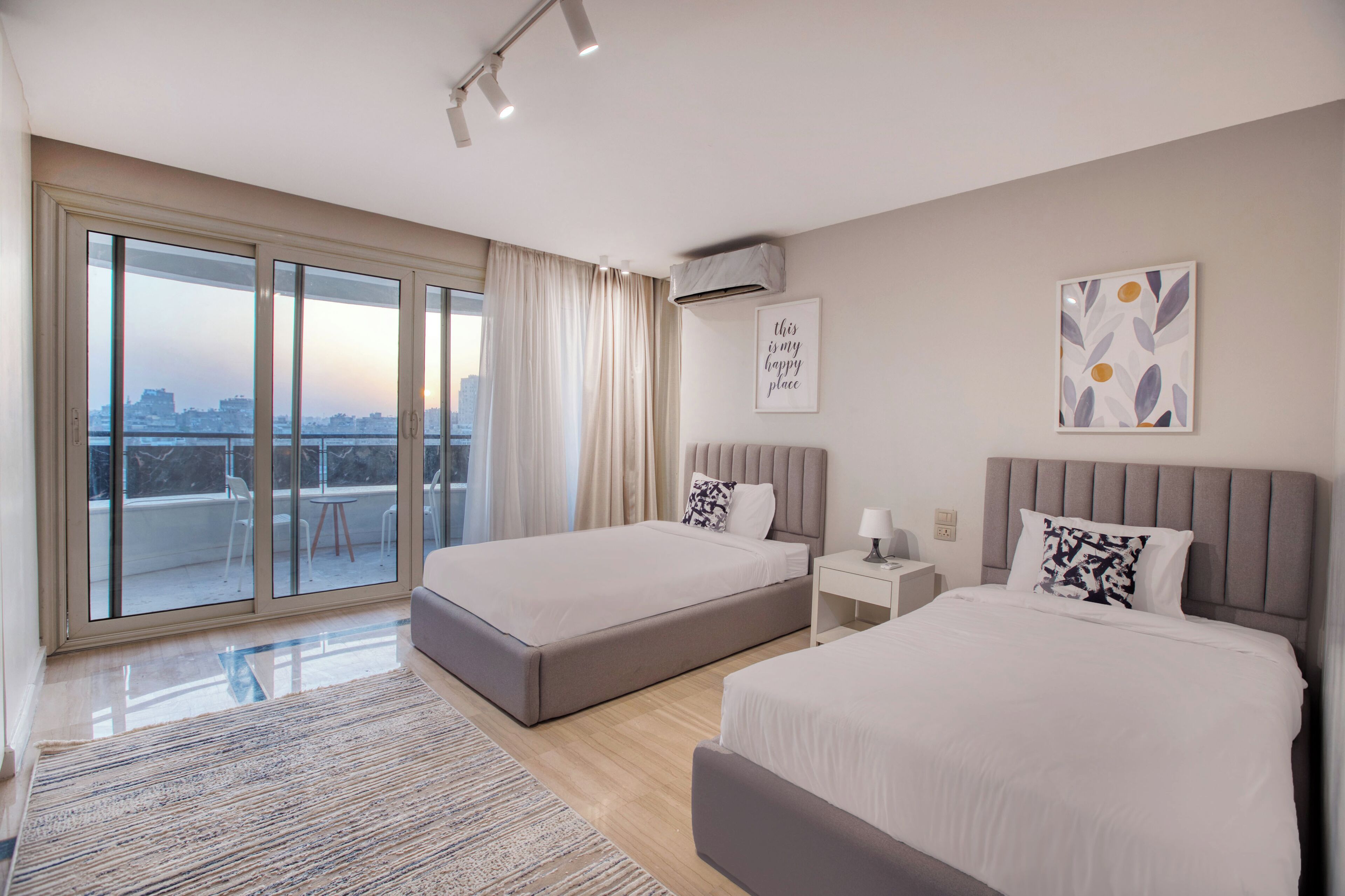 Ein modernes Hotelzimmer mit zwei Betten, einer großen Schiebetür zum Balkon, Blick auf die Stadt und einem gerahmten Zitat an der Wand.
