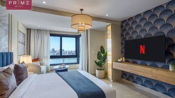 A modern hotel room with a large bed, a TV mounted on the wall, a city view, and a potted plant.