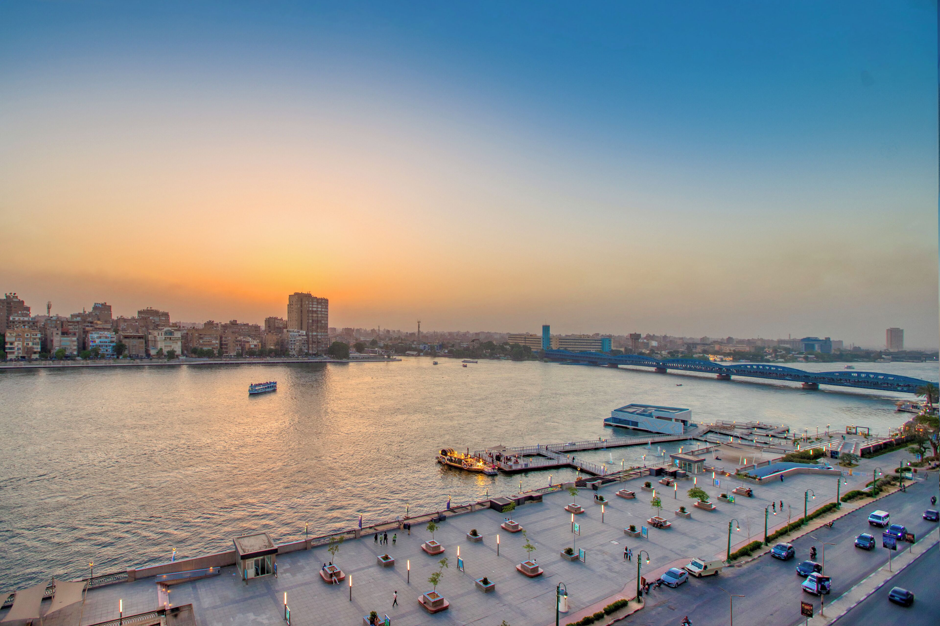 A waterfront cityscape with a bridge, a river, and a parking area with boats.