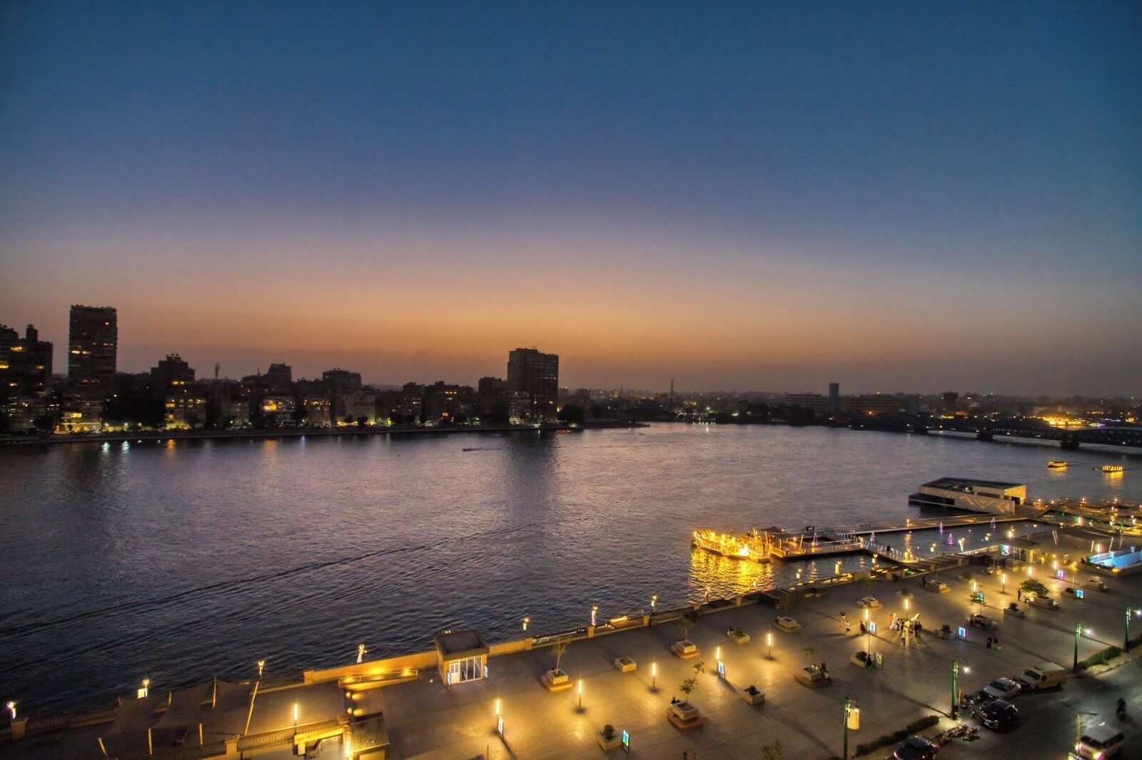 A waterfront cityscape at dusk with illuminated buildings and a dock area.