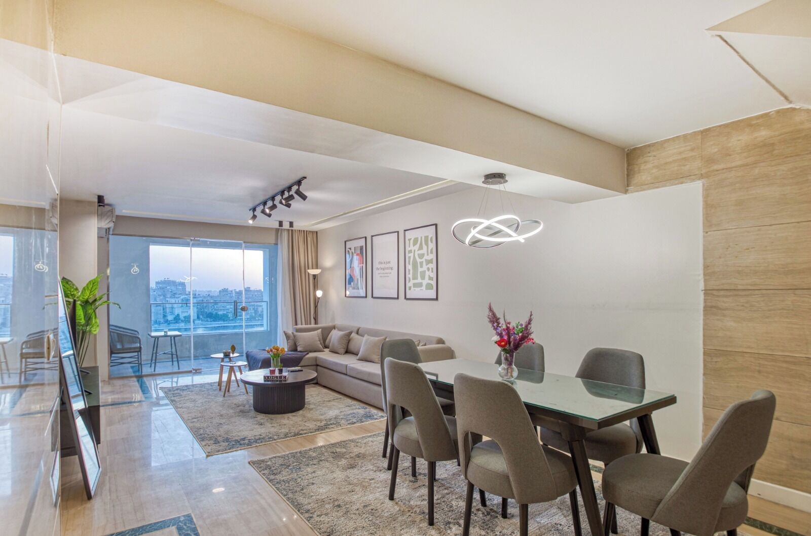A modern living room with a glass dining table, comfortable seating, a large window with a city view, and a stylish pendant light.