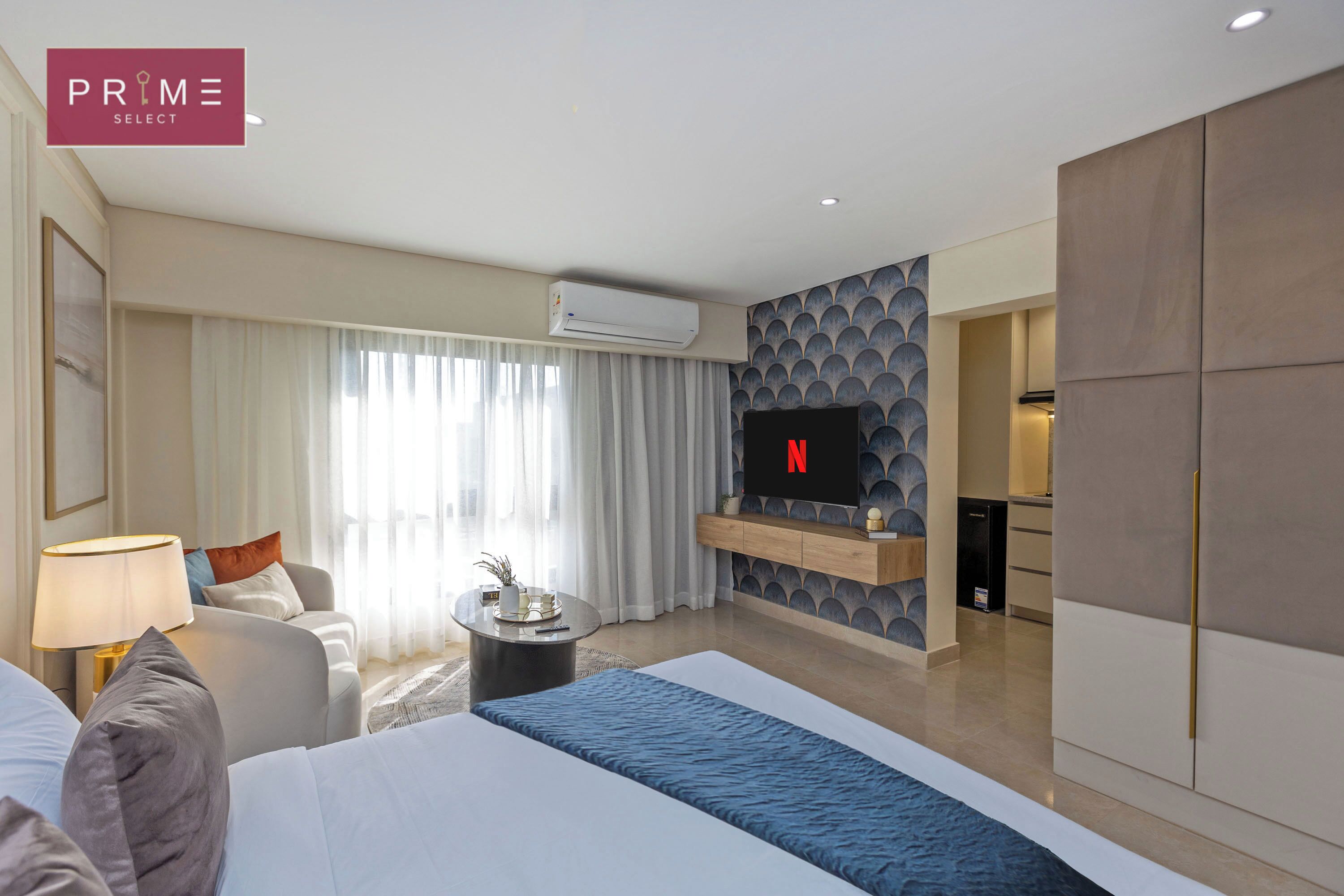 A modern hotel room with a large bed, a TV mounted on the wall, a desk with a lamp, and a view of the kitchen.