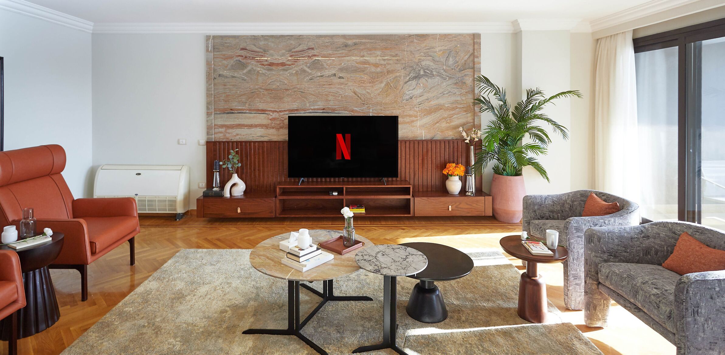 A modern living room with a flat-screen TV, red leather sofa, grey armchair, and a coffee table with books and a vase.