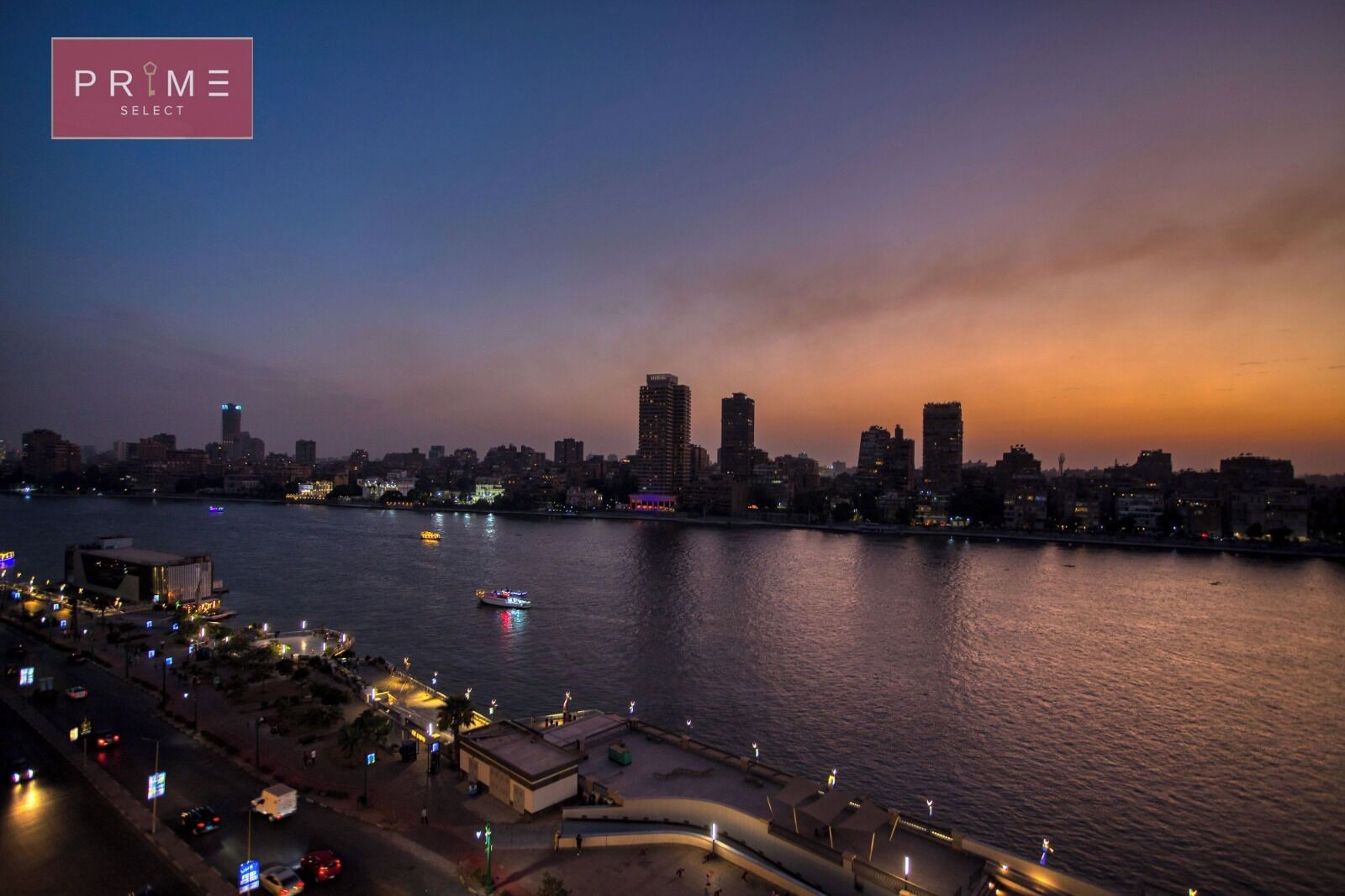 A nighttime cityscape with a river, buildings, and a waterfront promenade.