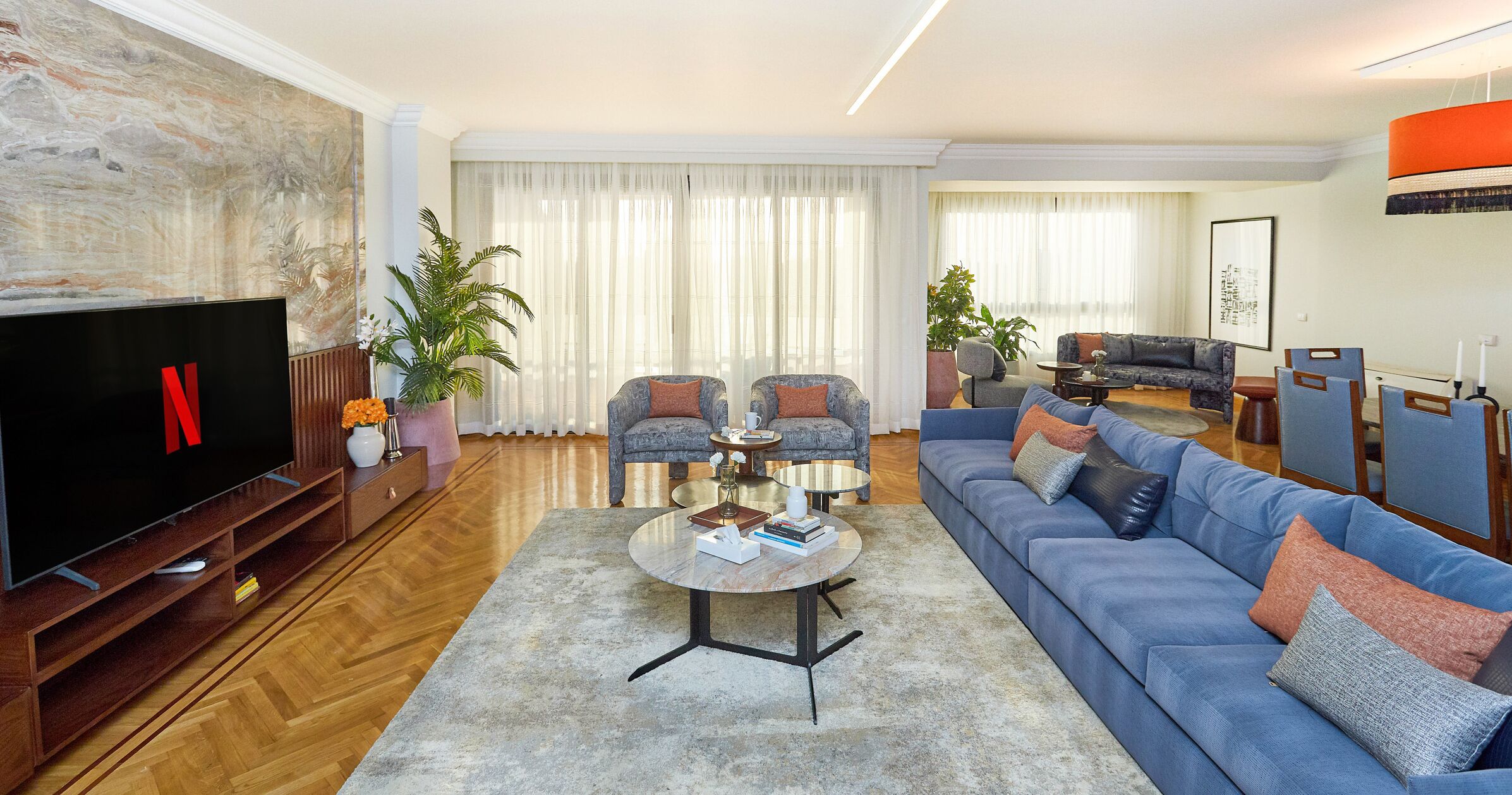 A modern living room with a large flat-screen TV, a blue sofa, two armchairs, a coffee table, and a large painting on the wall.