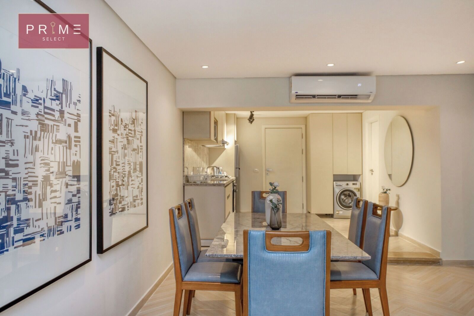 A modern dining area with a glass table and blue chairs, a kitchen with a washing machine, and a large framed cityscape artwork on the wall.