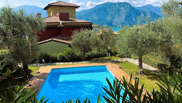 A pool surrounded by greenery, a building with a red roof, and mountains in the background. - LE VELE BETH'S HOUSE (Brenzone sul Garda)