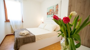 A bedroom with a bed, a bedside table with a lamp, a window with curtains, and a painting on the wall. - LE VELE BETH'S HOUSE (Brenzone sul Garda)