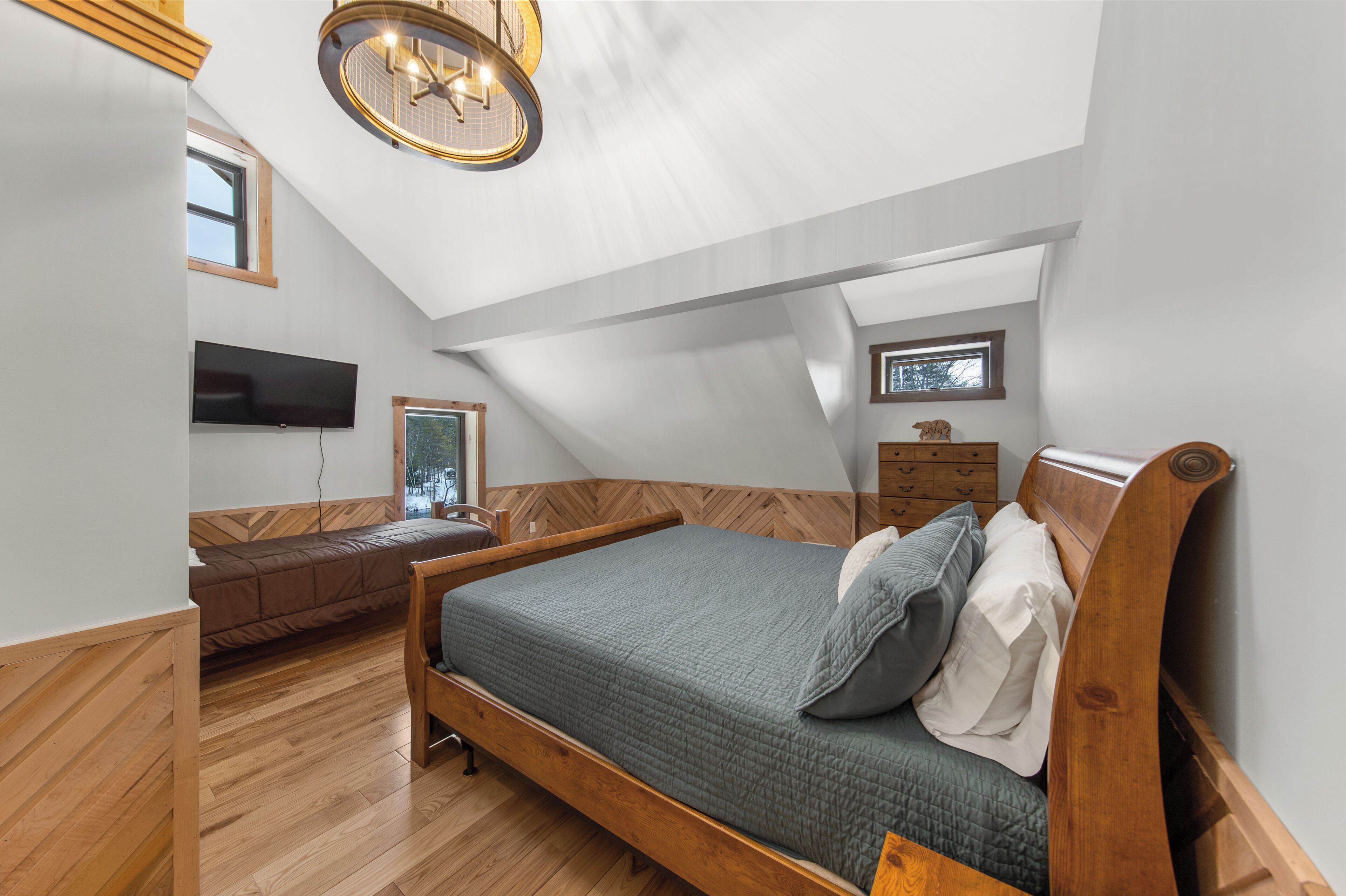 Une chambre avec un plafond en pente, un lit doté d’une tête de lit en bois, une commode, un téléviseur fixé au mur et une fenêtre.