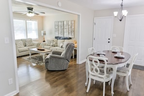 A spacious living room with a gray armchair, a wooden dining table with white chairs, and a ceiling fan. - The Merrimac House: Cute, Cozy and very Convenient to I-20 in Oxford, Al. (Oxford)