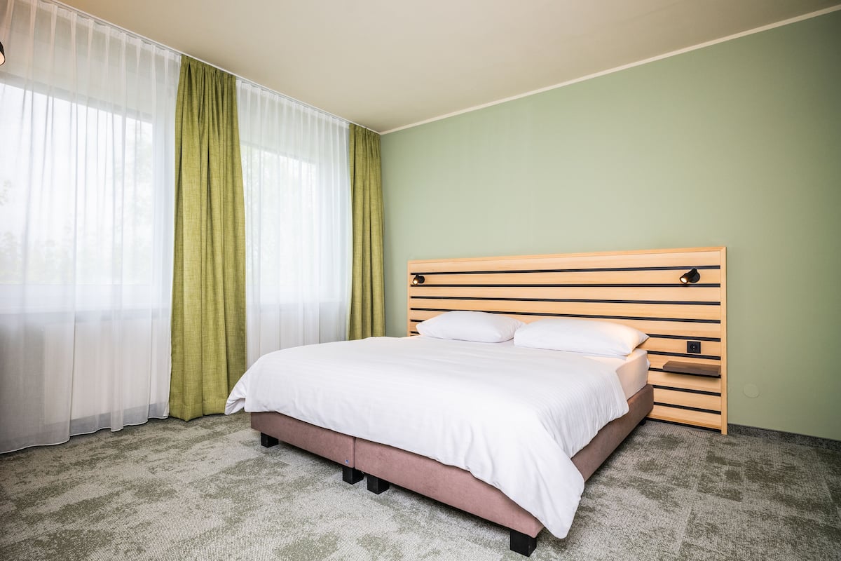 A neatly made bed in a room with a green wall, a wooden headboard, and sheer curtains.