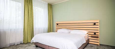 Una cama bien hecha en una habitación con pared verde, cabecera de madera y cortinas de gasa.