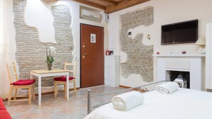 A hotel room with a stone wall, a fireplace, a TV, a bed, a table with chairs, and a red cushion.