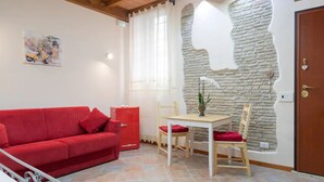A red sofa, a small table with two chairs, a decorative vase with white flowers, and a stone wall with a unique design.