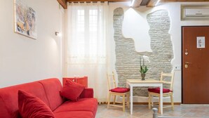 A red sofa with red cushions, a small wooden table with two chairs, a potted plant, and a stone wall.