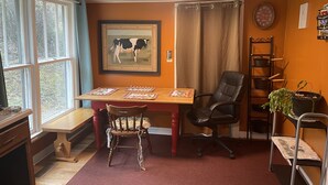 Een eetruimte met een houten tafel, twee stoelen, een bank, een schilderij van een koe in een lijst, een klok en een plank met keukenspullen.
