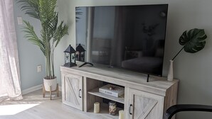 Ein Wohnzimmer mit Flachbildfernseher auf einer Holzkommode, einem Blumentopf und einem großen Fenster.