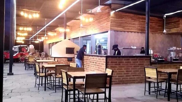 An outdoor cafe with wooden tables and chairs, a brick counter, and overhead lighting.