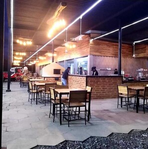 An outdoor cafe with wooden tables and chairs, a brick counter, and overhead lighting. - Hacienda San Agustin (Marinilla)
