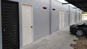 A row of industrial-style buildings with numbered doors, a covered parking area, and a black car parked on the right.