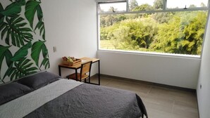 A bedroom with a bed, a small table, and a window with a view of greenery.