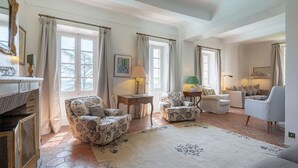 Tilava olohuone, jossa on takka, kaksi nojatuolia, sohva ja sohvapöytä. Suuret ikkunat, joissa on verhot, päästävät runsaasti luonnonvaloa huoneeseen.