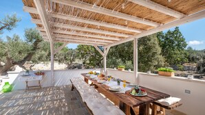 An outdoor dining area with a wooden pergola, stone flooring, and a view of olive trees and hills. - Charming Trullo with Heated pool and panoramic view (Cisternino)
