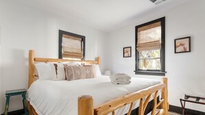 A wooden-framed bed with white bedding, two windows with bamboo blinds, and a small bedside table. - Stylish Monthly 1BR Downtown Rental (Charleston)