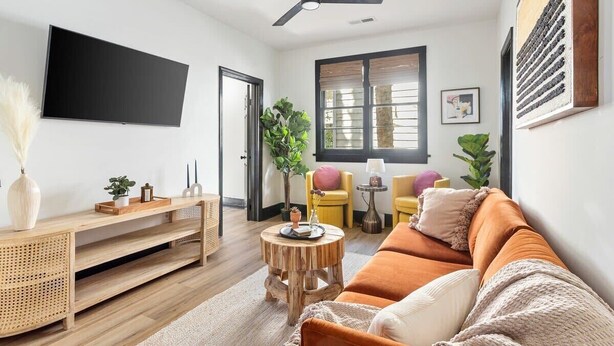 A modern living room with a flat-screen TV, a ceiling fan, a wooden coffee table, a sofa, and a window with blinds. - Stylish Monthly 1BR Downtown Rental (Charleston)